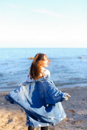 Smiling female walks along beach and enjoys nature, with broad smile on face jumping in place and raising hands upwards, squinting against bright sunlight, shielding eyes with hands. European-looking girl with medium-length blond hair dressed in black panの写真素材