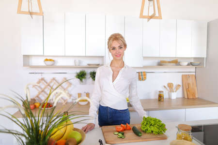 Slim, sweet girl looks away and with beautiful smile on face poses, spreads vegetables on cutting board, standing in middle of stylish and light kitchen equipped with modern technology.の写真素材