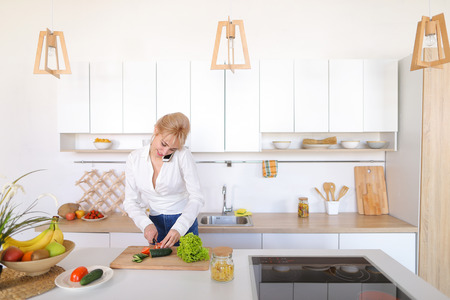 Stylish female housewife communicates on gadget with friend and stands with back to camera, turns and discusses with girlfriend latest gossip on honeycomb, slicers vegetable salad of red tomato and fresh green cucumber on cutting wooden board amidst styliの写真素材
