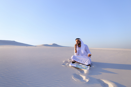 Handsome Arab businessman, talking on cell phone with companion about future construction and general business, looks at drawing and makes adjustments with ballpoint pen, sitting on white clean sand in open air desert on summer day. Swarthy Muslim with shの写真素材