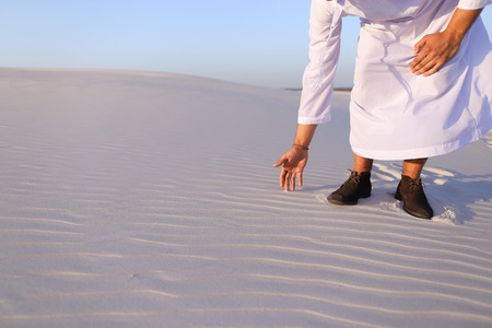 Happy young Emirati Sheikh bends over and takes white sand in handsの写真素材