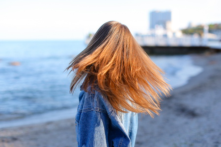 Charming girl resting and with beautiful smile on face posing in camera, laughing and enjoying beautiful day on shore of blue sea. Woman of European appearance with blond mid-length hair dressed in blue denim jacket and stands with back to camera lens agaの写真素材
