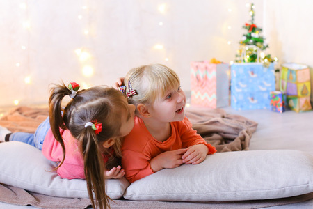 Lovely little girls look at camera and at each other, laugh and smile, play games and communicate, share secrets and lie on soft cushions in bright room with New Years small Christmas tree and garland on wall. Blond girl of European appearance with tails の写真素材