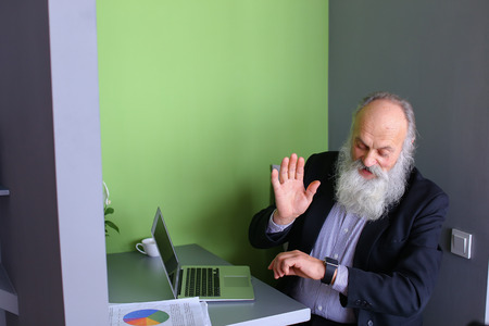 Oldie modern man study new and modern technologies, learn to work with headsets and, sitting and working on laptop in cozy computer cafe on sunny day. Elderly man with long gray beard of European appearance dressed in black classic suit and white shirt inの写真素材