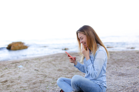 Cute girl holding cell phone and leads in social network or post photos, laughing and smiling, sharing impressions of beauty of sea, sitting on sandy beach on backdrop of sea and sky on clear evening . Young European-looking woman with long blond hair dreの写真素材