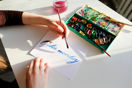 Creative woman holds thin wooden brush in hands and writes in beautifully italic typeface text from letters Cyber Monday on white sheet of paper in blue colors, sitting at white table on which there watercolor and glass jar with water in art studio. Europの写真素材