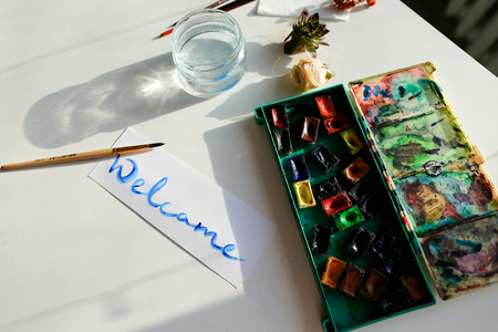 Greeting card with blue calling inscription welcome, painted with watercolors on white rectangular sheet of paper, which lies on light table. Concept of calligraphy and lettering, fine arts and bright colors, beautiful writing, materials and tools, craftsmanship and hobbies, sales and advertising.の写真素材