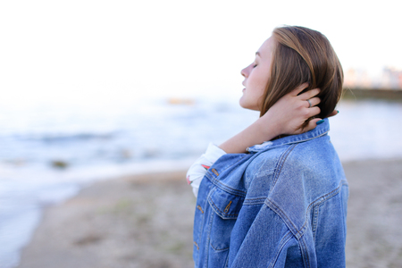 Beautiful young woman with necessary smile looks afar at sea waves, closes eyes with pleasure and listens to sound of surf, poses and warms from cool wind from sea on clear cool evening. Woman of European appearance with blond mid-length hair dressed in black pants, white T-shirt with bright pictures on top of which blue denim jacket stands against blue sea and sky. Concept beautiful and happy people, unity with nature, joyful and cheerful mood, fashion and style, natural and natural beauty, rest by sea and enjoyment of sea breeze, summer heat in resort city, advertising of women's clothing or cosmetics.の写真素材