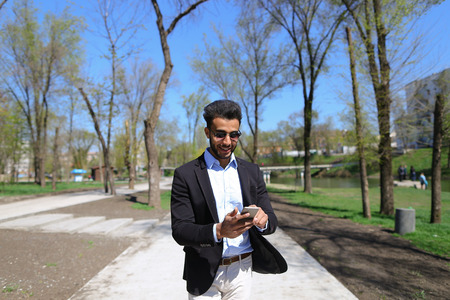 Top-down shooting, boy stands by wooden building and texting massage on phone, wears sunglasses. Guy has beard, short hair and dimples on cheeks. Man dressed in black jacket, white jeans and blue shirt, brown shoes. Concept of new brand clothes modern collections of shoes.の写真素材