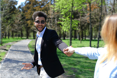 Gay leading aunt for walk in park. Man has beard, short hair and dimples on cheeks. Boy dressed in black jacket, brown belt on white jeans and blue shirt, wears sunglasses. Concept of places for relaxing family time and day off.の写真素材