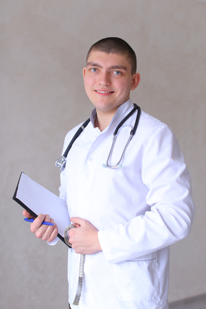 Handsome male specialized doctor poses on camera in good mood and smiles, loves work and gives advice with folder and centimeter tape in hands, and stethoscope on neck and  standing in bright medical cabinet. Guy physician  European-looking with short haircut dressed in white medical uniform. Concept of people and professions, consultative reception of doctor, medicine and pharmacology, treatment of diseases by drugs, oath of Hippocrates, medical equipment and devices.の写真素材