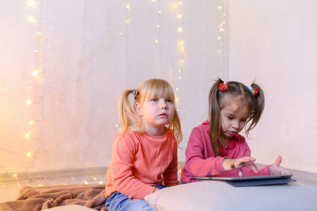 Cute little girls play in computer games on device or watch cartoons, press something on touch screen of tablet, smile and communicate with each other, sitting on rug in bright room with flickering New Years garland in evening. Blond girl of European appearance with tails on sides dressed in orange sweatshirt with long sleeves and blue jeans, small female brunette with two tails on each side dressed in raspberry sweater and blue jeans. Concept of happy and carefree childhood, modern technologies and gadgets, bright and unforgettable New Years childrens emotions, Christmas gifts and Santa Claus, childrens entertainment and fun games, decor and bright festive interior.の写真素材