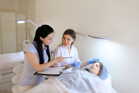 Specialist in microblading, beautiful girl shares her secrets of working with eyebrow correction tools and shows basic techniques to female assistant on example of woman lying on couch in well-equipped beauty salon. Young girl future master of eyebrow tattoo records information on piece of paper and closely watches. Woman of European appearance with light brown hair dressed in white medical gown and blue rubber gloves, European-looking woman with long black hair dressed in blue polo shirt, European-looking girl with fair-haired hair lying on couch. Concept of cosmetology procedures and microblading, beautiful and educated people in profession, medical special clothes, regular customers and receptionist, professionally equipped for salon, beauty and appearance, training and transfer of experience to newly masters of tattoo.の写真素材