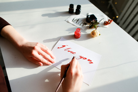 Female master of calligraphy and lettering creates red ink and pen fountain with wonderful love valentines with text I love you on day of all lovers on light table with accessories for lettering, small jars with colorful mascara and decorations in art studio. Concept of beautiful letter, calligraphy and lettering, materials and tools, craftsmanship and hobbies, recognition on Valentines Day, fine art and bright colors.の写真素材