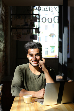 Dark-skinned young man sits in caf by table with laptop, drinks coffee and dreamily looks out the window. Bearded handsome man with dark eyes take few sips of coffee and smile. Cheerful guy with perfect smile look straight to camera. Concept of socials works or for advertising caf .の写真素材