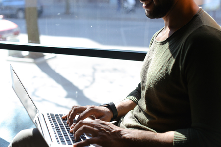 Journalist using laptop and sits near window. Boy dressed in white jeans, brown shoes and khaki pullover. Guy has beard and black hair. Concept of modern technologies working at home.の写真素材