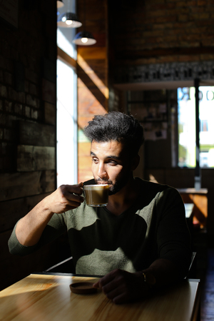 Boy sits in comfortable cafÃÂ© and drinks coffee. Smiling man has dimples, beard, full lips and black hair. Guy dressed in khaki pullover. Concept of comfortable places for wedding tasty coffee.の写真素材