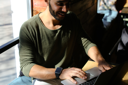 Insurance agent checks mail and clicking on keyboard. Boy dressed in khaki pullover. Attractive man has dimples, beard, full lips and black hair, long fingers. Concept of modern technologies and work in Internet online wifi.の写真素材