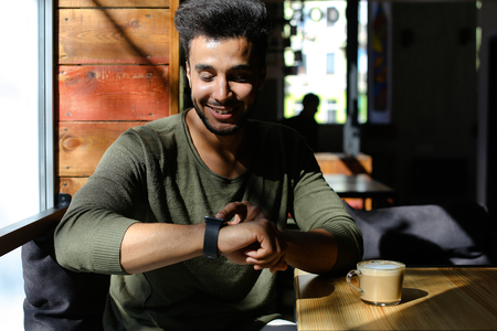 Man sitting and puts hand on table. Boy has dimples, beard, full lips and black hair. Male dressed in khaki pullover and white jeans. Concept of new modern places for resting comfortable.の写真素材
