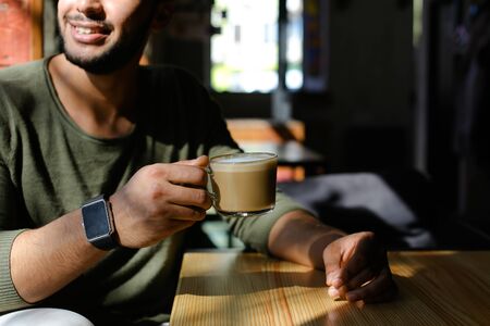 Handsome man sits near window and drinks coffee. Guy has short hair, beard and wears smart watch on...