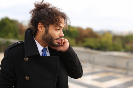 Half Italian and Nigerian man making call with modern smartphone close to Eiffel Tower in slow motion. Young male person has black curly hair, nice smile and earing, wears black coat. Concept of new technologies for young businessman, apps for talking by Internet always in touch.の写真素材