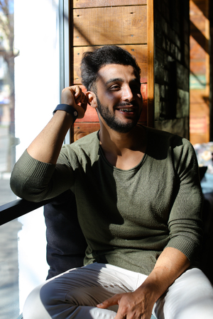 Man sits in cafe and looking in window and smiling with dimples. Person dressed in khaki pullover. Handsome male has beard, full lips and black hair.の写真素材