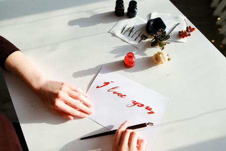 Female master of calligraphy and lettering creates red ink and pen fountain with wonderful love valentines with text I love you on day of all lovers on light table with accessories for lettering, small jars with colorful mascara and decorations in art studio.の写真素材
