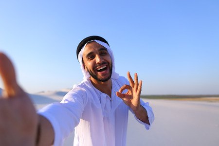 Close-up portrait of attractive Muslim guy holding camera with hand, aiming at himself and smiling with broad smile.の写真素材