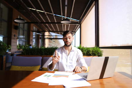 European businessman working at table with laptop and reading documents. Successful man dressed in white shirt has beard. Concept of using fast internet and modern technologies for biz.の写真素材
