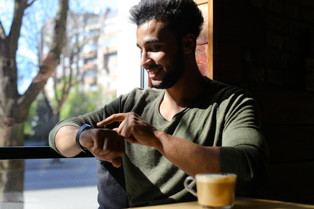 Arab guy sits in caf and texting e-mail by smart watch. Happy boy has dimples, beard, full lips and black hair. Person dressed in khaki pullover. Concept of modern technologies and good atmosphere.の写真素材