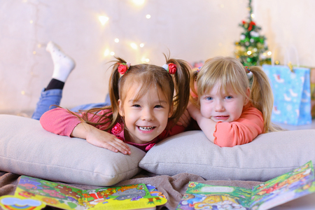 Funny little girls friends read childrens bright book and look at pictures, laugh and smile at each other, play games and talk, pose on camera and lie on soft cushions in bright room with New Years small Christmas tree and garland on wall. Blond girl of European appearance with tails on sides dressed in orange sweatshirt with long sleeves and blue jeans, small female brunette with two tails on each side dressed in raspberry sweater and blue jeans. Concept of happy and carefree childhood, bright and unforgettable New Years childrens emotions, Christmas gifts and Santa Claus, childrens entertainment and fun games, decor and bright festive interior.の写真素材