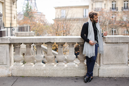 Mulatto tourist raving with white in ear headphones and smartphone near Eiffel tower. Jocund guy visiting Paris and enjoying with popular songs. Concept of using modern gadgets for entertainment and trip to France.の写真素材