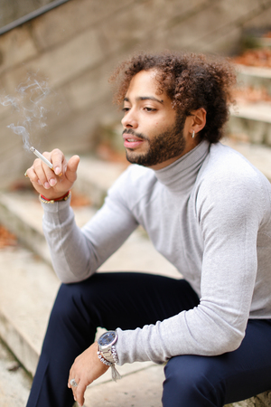 Upset mulatto male person sitting o steps with cigarette and smoking.  Young guy dressed in grey turtleneck sweater thinking about problems decisions and wears red string. Concept of autumn depression and bad habits.の写真素材