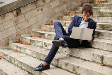 Mix blood student resting after classes with laptop, lying on steps. Young guy chatting with friend outside. Concept of lounging with modern gadget outside and enjoying Internet communication.の写真素材