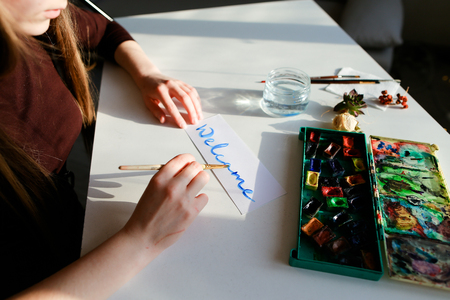 Female calligrapher holds brush in hand and writes welcome note on white background with watercolor blue colorsの写真素材