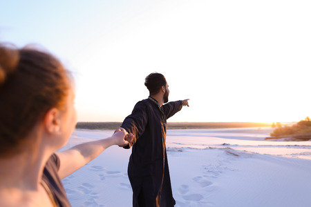 Pair of young lovers, male Muslim and woman of European appearance, look into each others eyes and hold hands, walking along wide, bottomless sandy desert. guy and girl are smiling and laughing in open air at sunset.の写真素材