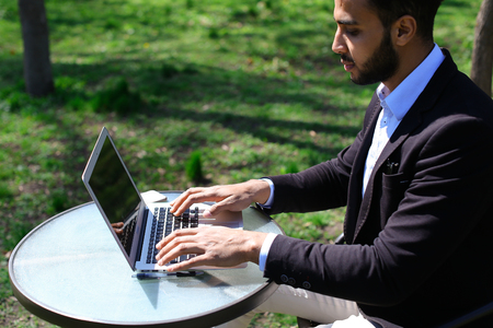 Handsome person using laptop and clicking keyboard, putting hands up. Male has beard, short hair and dimples on cheeks. Guy dressed in black jacket and blue shirt. Concept of modern technologies new apps on laptop online selling.の写真素材