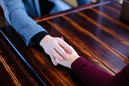 Boyfriend want to make girlfriend offer of hand and heart in favorite cafe, couple waiting for order and fooling. Young fair-haired woman with neat red manicure and guy in blue jacket sitting near wooden table. Concept of cozy luncheonette and nice atmosphere, quiet coffee house for loversの写真素材