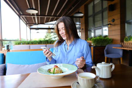Beautiful female person typing messages by smartphone at cafe and eating dessert with cup of tea  . Young woman resting at restaurant and chatting with boyfriend. Concept of communication, gadgets and having sweet lunch.の写真素材