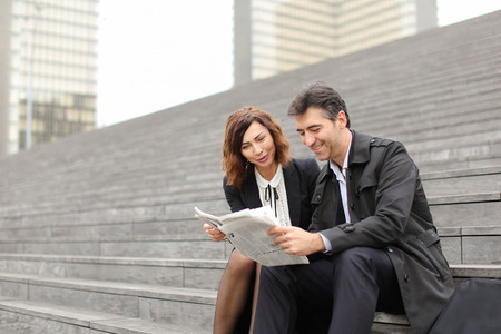 engineers male and female find article about company in newspaper, colleagues sitting on stairs reading. Americans wearing business clothes smiling communicating. Concept of fashionable clothes, woman and man outfits or mass media.の写真素材
