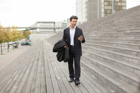 business man using smartphone walk, husband watching photos of wife in working trip. Middle-aged American wearing black jacket and white shirt smiling. Concept of innovative technologies and gadgets, designer clothing or fashionable man outfits.の写真素材