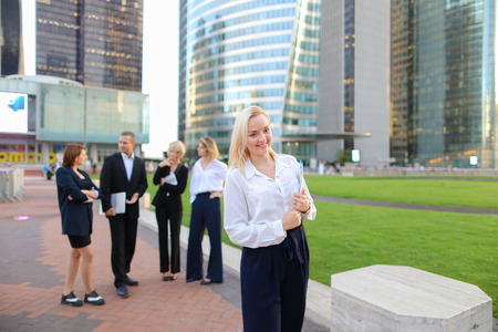 Close up face blonde secretary looking at camera and smiling with blue case in   and talking partners background. Concept of office assistant with documents outside and business employees. Beautiful blonde girl with casual make up and cute smile.の写真素材