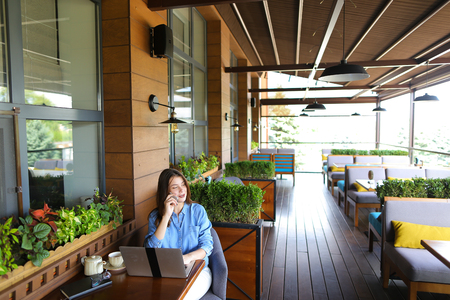 Female student chatting with friends by laptop at cafe. Young girl dressed in jeans shirt sitting near window with room plants. Concept of communicating with close people and modern technologies.の写真素材
