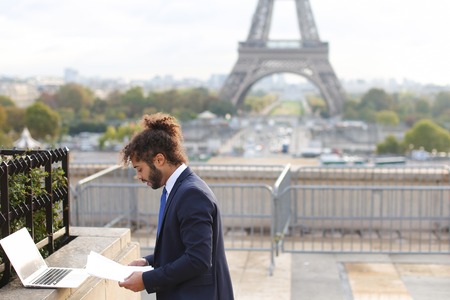 Mulatto man using laptop on open air around Eiffel Tower in slow motion. Handsome cute boy wears in suite with blue tie, has black curly hair and beard. Concept of modern technologies for work using new apps.の写真素材