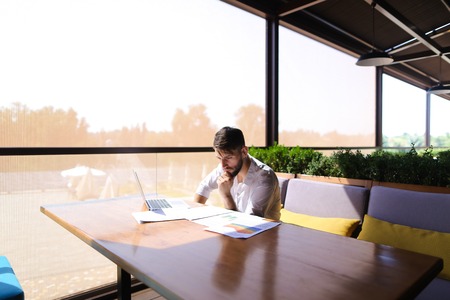European businessman working at table with laptop and reading documents. Successful man dressed in white shirt has beard. Concept of using fast internet and modern technologies for biz.の写真素材