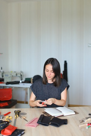 Female slicing leather using special knife, ruler and scissors, young lady recently got job in atelier. Craftswoman with neat red manicure standing near wooden table with tools in well-lit workshop and preparing materials for making shoes. Concept of accuracy, handicraft or fine work.の写真素材
