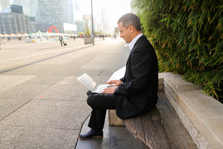 Businessman typing by laptop outside near green plant in   and looking at watch. Concept of ecology thinking for business. Successful man dressed in black suit working with computer outdoors.の写真素材
