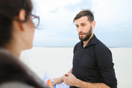 race driver meet with journalist female to tell about hobby, people shake hands at end of conversation. Lady with ponytail in white blouse beige skirt holding planchette talking with bearded male in black shirt trousers. Concept of collecting information for article, fashionable clothes or stylish man and woman outfits.の写真素材
