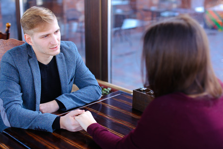 Boyfriend want to make girlfriend offer of hand and heart in favorite cafe, couple waiting for order and fooling. Young fair-haired woman with neat red manicure and guy in blue jacket sitting near wooden table. Concept of cozy luncheonette and nice atmosphere, quiet coffee house for loversの写真素材