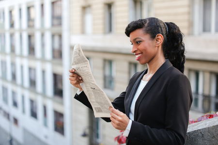 Half African girl reading newspaper near high building with close up of face. Concept of learning new information from public press.の写真素材